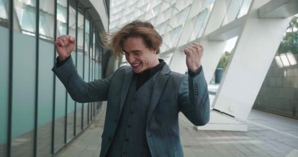 Excited businessman rejoices at victory, clenches his fists, shows yes. Satisfied male entrepreneur celebrates successful win while standing outdoors near modern office building — Video Stock