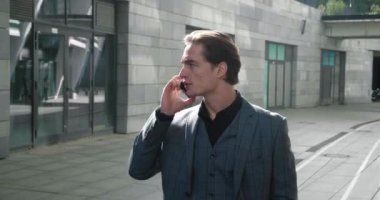 İş merkezinde yürürken resmi takım elbiseli genç bir iş adamı telefonda konuşuyor. Gülümseyen erkek girişimci işe giderken cep telefonuyla konuşuyor.