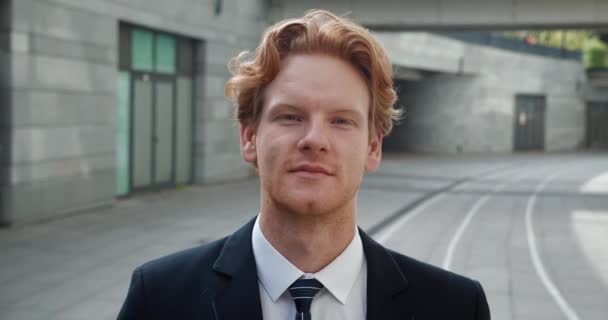 Confident young redhead businessman looks at the camera and smile while standing in the business district near office building. Wearing formal suit. Close-up portrait. — Stock Video