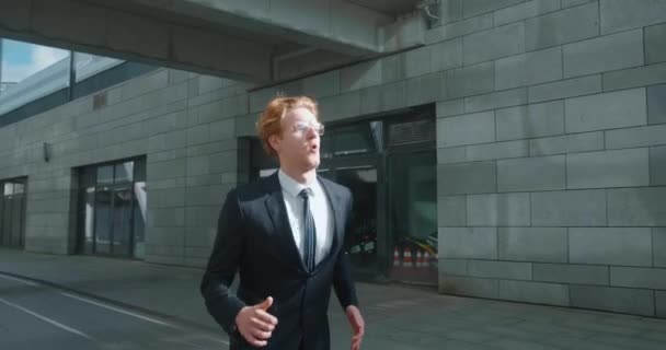 Young businessman runs down city street. Business man late for meeting. Successful man in formal suit jogging near modern office buildings. Red-haired male manager in hurry to appointment. — стоковое видео