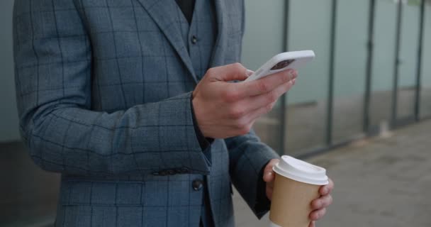 Close up of businessmans hand using mobile phone, surfing internet with smartphone and holding coffee, standing outside near office building — Vídeo de Stock