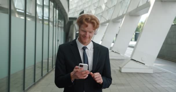 Retrato de un alegre hombre de negocios pelirrojo con auriculares inalámbricos que escucha música usando un teléfono inteligente y baila divertido, parado al aire libre cerca de un edificio de oficinas — Vídeo de stock