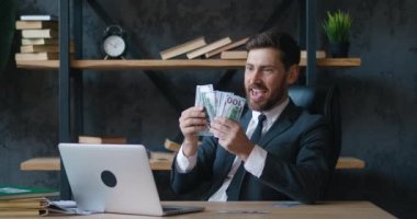 Cheerful bearded businessman in formal suit throwing money dollar bills having fun at desk in modern office alone enjoying wealth. Rich people, lifestyle and business concept.
