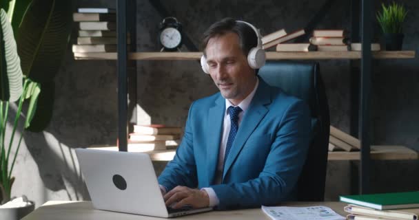 Portrait of mature male businessman enjoying music with headphones while working with laptop at modern office. — Video