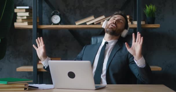 Feliz barbudo maduro hombre de negocios escuchando música en los auriculares mientras trabaja en el ordenador portátil en la oficina — Vídeos de Stock