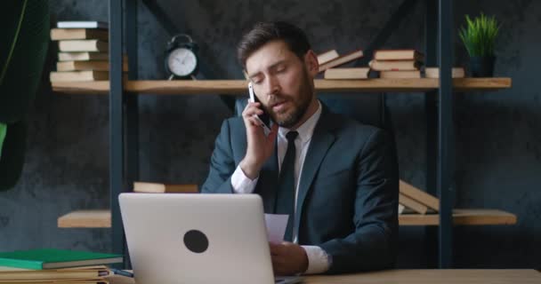 現代のオフィスに座ってラップトップコンピュータで働いている間、若いひげを生やしたビジネスマンが携帯電話で彼のビジネスについて通信します。 — ストック動画