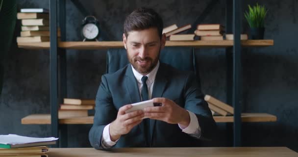 興奮したビジネスマンは、スマートフォンを保持します喜び男性の勝者は、現代のオフィスに座っている間、携帯電話で読み取り良いニュースを受け取るモバイルオンライン入札ゲームの勝利の成功を祝います — ストック動画