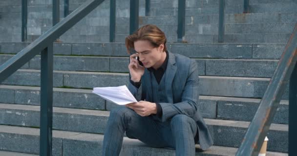 Busy businessman with papers in his hands sitting on stairs near office center building and talking on phone. Male executive having phone call sitting on staircase outdoors. Concept of workload — Stockvideo