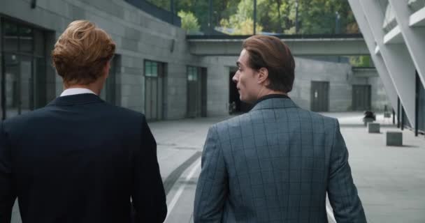 Rear back view of two business men colleagues in formal suits chatting on the way to work. Two males employees managers talking while walking at downtown. — Stock videók