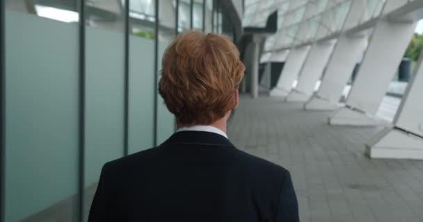 Back view of ambitious young red-haired man entrepreneur walks along the city street downtown. Rear view of confident businessman wearing suit, commuting to work at business district — Stockvideo