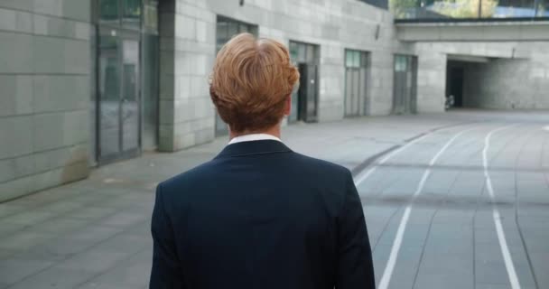 Rear back view of ambitious young man entrepreneur walks along the city street downtown. Confident red-haired businessman wearing suit, commuting to work. Steadicam shot, slow motion — стоковое видео