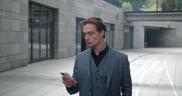 Young businessman using smartphone texting walking along office building checking email messages online successful male executive going to work — Stok video