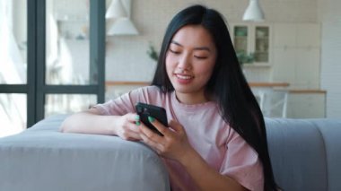 Front view of a serene happy dark-haired Asian woman using her smartphone for texting, browsing mobile apps, swipping, shopping and booking. The place of digital electronic devices in everyday life