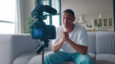 Millennial african blogger or vlogger man filming new vlog video with professional camera, mounted on tripod sitting on sofa at home.
