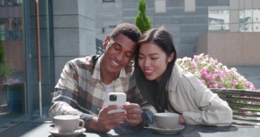 Young couple sitting together in the outdoor cafe on beautiful sunny day at romantic date, talking, looking at the screen of cell phone in the hands of handsome guy, swiping, surfing on web pages