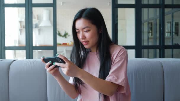 Excited young Asian woman holding mobile phone and playing virtual computer games, enjoying victory, typing and looking at touch screen, smiling toothy smile. Computer virtual games addiction — стоковое видео