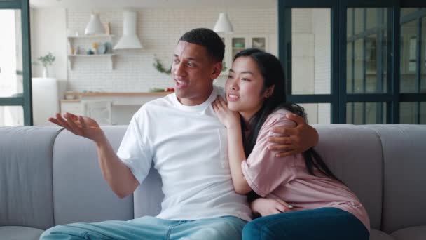 Young asian woman leaning on the shoulder of her african american boyfriend, relaxing on the couch at home on her day off, discussing together, looking away, enjoying spending time together indoors — Stock Video