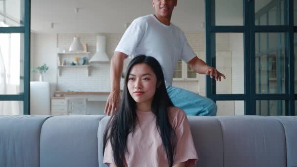 Cheerful and handsome young mixed race African American man coming up from behind and covering eyes of his Asian girlfriend sitting alone on sofa in modern smart apartment, making her surprise. — Stock Video