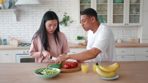 Overjoyed mixed race African American man and Asian woman enjoying cooking healthy tasty vegan salad in kitchen together. Smiling young girl cutting cucumber on a wooden desk in own cozy house.