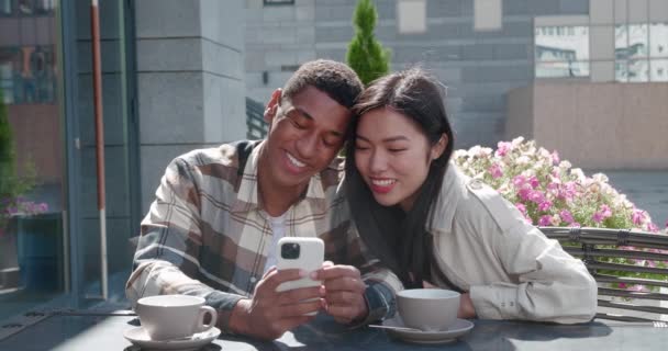Young couple sitting together in the outdoor cafe on beautiful sunny day at romantic date, talking, looking at the screen of cell phone in the hands of handsome guy, swiping, surfing on web pages — Video Stock