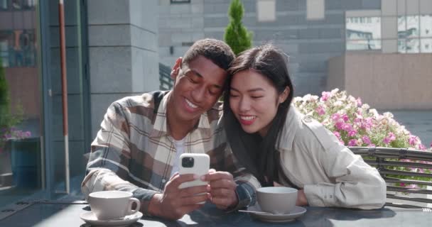 Pareja joven sentados juntos en la cafetería al aire libre en un hermoso día soleado en una cita romántica, hablando, mirando la pantalla del teléfono celular en manos de un chico guapo, deslizando, navegando en las páginas web — Vídeos de Stock