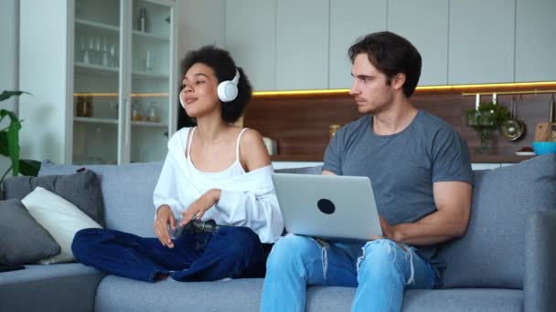 Attractive African woman in headphones dancing singing sitting on the couch next to her boyfriend. The man concentrated on working at the laptop, nervously looks in her direction in bewilderment. — Vídeo de Stock