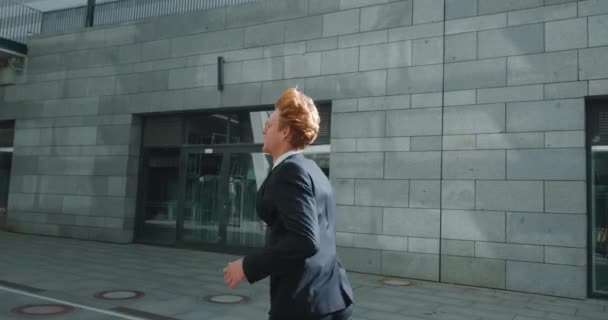 Young businessman runs down city street. Business man late for meeting. Successful man in formal suit jogging near modern office buildings. Red-haired male manager in hurry to appointment. — Stock Video