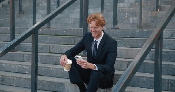 Happy young businessman wearing glasses sits outdoors on steps of office building, using smartphone and drinking coffee. Red-haired male employee in formal suit with mobilephone during coffee break — kuvapankkivideo