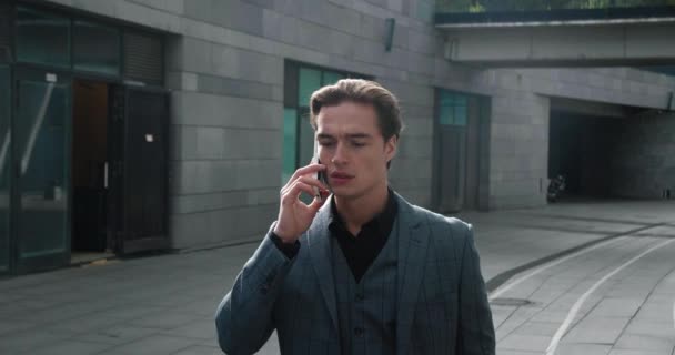 Confident male businessman in formal suit emotionally talking mobile phone while walking outside at downtown near office buildings in slow motion — 图库视频影像