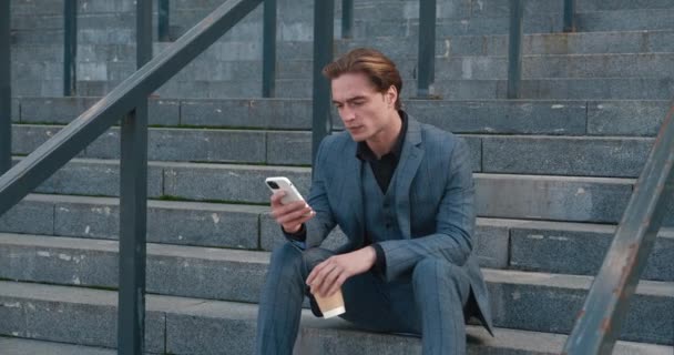 Handsome man in business suit sitting on stairs at coffee break and typing on smartphone, near office building, chatting with friends or doing shopping — Stock video