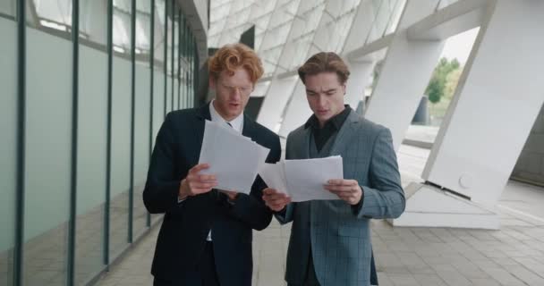 Caucasian two handsome stylish businessmen standing outdoor and talking. Man showing paper documents to male colleague, discussing a business plan, standing outdoors at business district downtown — Video