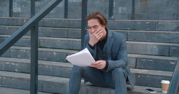 Sad young businessman in a suit with business papers in his hands sits on the steps of an office building. The concept of workload and burnout — Stok video