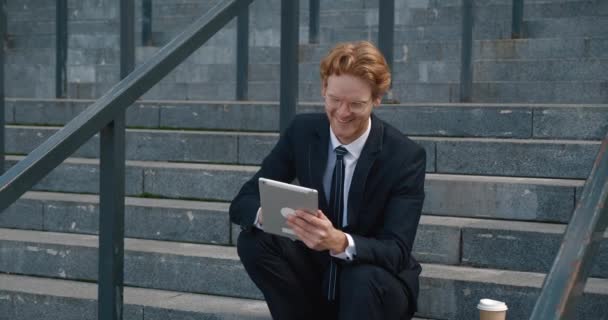 Redhead happy man in formal suit sitting at stairs near modern office building and using tablet pc — Video Stock