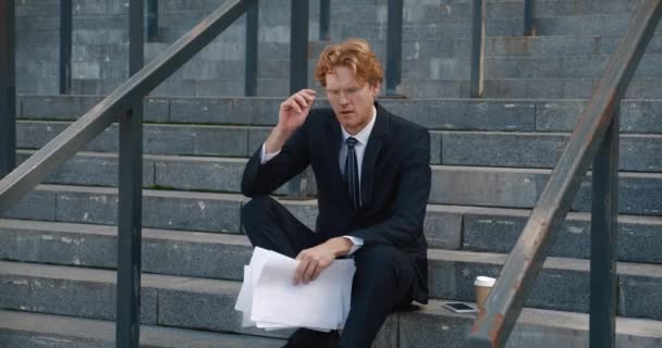 Stressed businessman with business papers in his hands sits on the steps of an office building. Sad overworked redhead man manager, takes off glasses, putting hands to the head suffering from headache — Videoclip de stoc