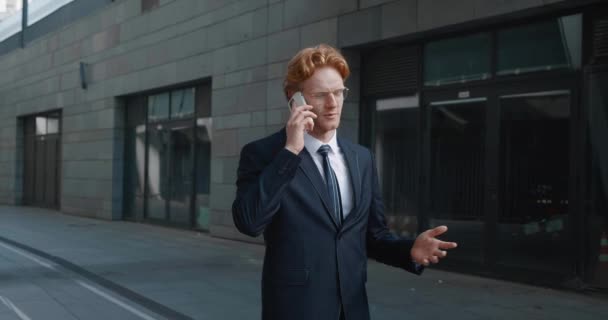 Молодий бізнесмен в офіційному костюмі говорить по телефону під час прогулянки в центрі міста в діловому районі. Усміхнений рудий чоловік-підприємець розмовляє на мобільному телефоні під час роботи — стокове відео