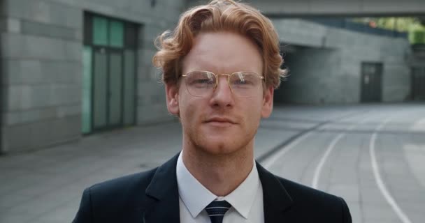 Confident young redhead businessman wearing glasses and looks at the camera while standing in the business district near office building. Wearing formal suit. Close-up portrait. — Stock video