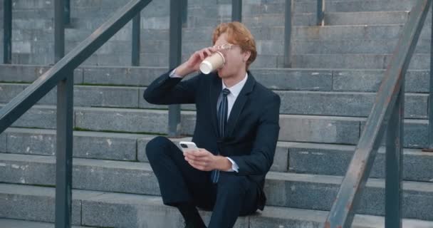 Усміхаючись молодий бізнесмен сидить на вулиці на сходах офісної будівлі, використовує смартфон і п'є каву. Рудий чоловік у діловому костюмі використовує мобільний телефон під час перерви на каву — стокове відео