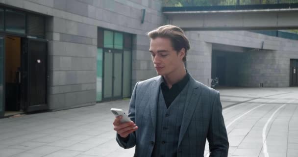 Confident handsome business man in formal suit checking social networks on his smart phone while walking along business buildings at downtown. — Stok video