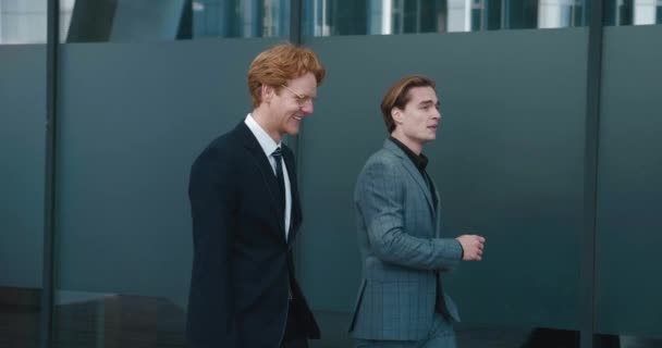 Side view of two smiling businessmen discussing startup while walking outside office at break. Males financial investors are talking outdoors. Two colleagues communicate on the way to work — 图库视频影像