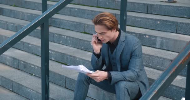 Businessman with business papers in his hands sitting on stairs near office center building and talking on phone. Male executive having phone call sitting on staircase outdoors. Concept of workload — Vídeos de Stock