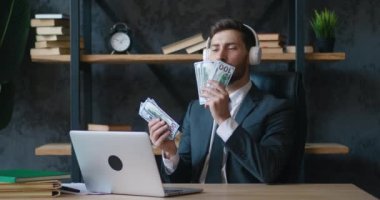 Mutlu sakallı genç iş adamı kulaklıkla müzik dinliyor ve para saymayı seviyor. Kolay kazanç konseri. 4K