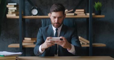 Akıllı telefonuyla oynayan sakallı bir iş adamı modern ofiste masa başında otururken yumruğunu sıkarak yarış oyunu kazanıyor. 4K