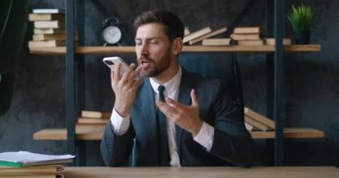 Ofiste oturan sakallı genç işadamı hoparlörden arıyor. Resmi takım elbiseli neşeli erkek iş yerindeki sesli mesajları kaydetmek için cep telefonu kullanıyor. 4K