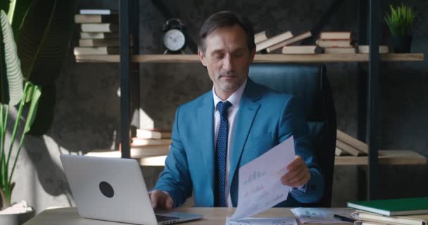 Portrait of mature businessman in formal suit. Man entrepreneur sitting at the table in front of laptop and working with papers in modern office — Video