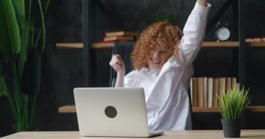 Heyecanlı kızıl saçlı genç iş kadını modern ofiste dizüstü bilgisayarla masada oturuyor. Harika haberler alıyor ve çok mutlu. Evet işareti gösteriliyor
