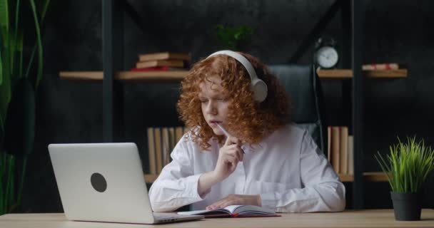 Kızıl saçlı, kulaklık eğitimi alan genç bir bayan öğrenci. İnternetteki öğretmen ile video konferansında sohbet ediyor. Video konferansını arıyor. — Stok video