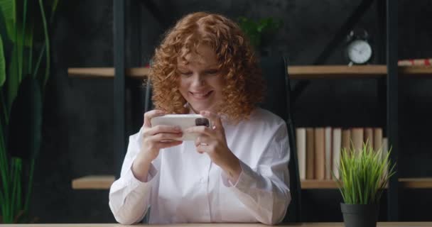 Joven mujer pelirroja positiva concentrada sentada en el escritorio de la oficina, jugando juegos de carreras en línea por teléfono móvil hacer gesto ganador — Vídeos de Stock