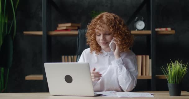 Щаслива молода руда бізнес-леді професійно розмовляє по телефону під час роботи з ноутбуком, сидячи за офісним столом, дивлячись на бізнес-документи — стокове відео