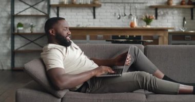 Erkek girişimci evde kanepede yatarken bilgisayarını uzaktan video görüşmesi için kullanır. Afrikalı serbest çalışan oturma odasındaki kanepede video konferansı yapıyor..