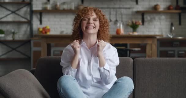 Потрясенная молодая рыжая женщина с вьющимися волосами, показывающая жест "да", сидя дома. Молодая женщина празднует победу. Концепция выигрыша в лотерею. — стоковое видео